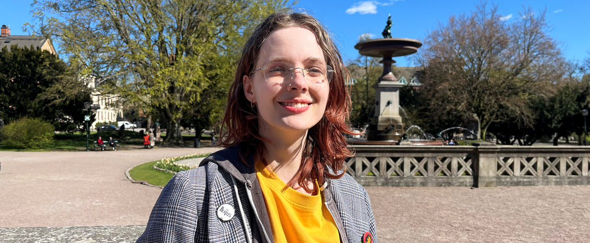 Olivia Persson. En person i gul t-shirt och rutigt kavaj står i en park och ler.